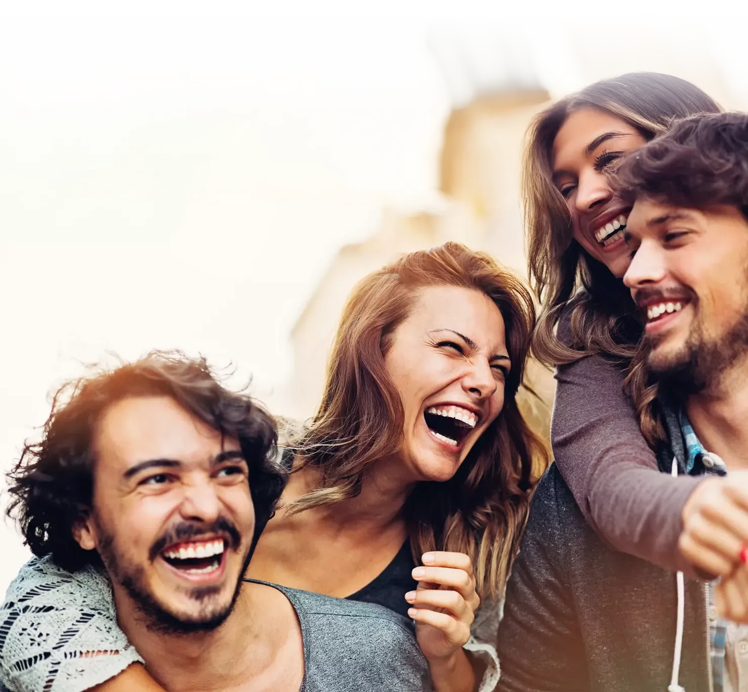 Vier junge Freunde lachen ausgelassen im Freien und teilen sichtlich einen fröhlichen Moment. Sie stehen eng beieinander, strahlen vor Freude und wirken unbeschwert. Die warmen Farbtöne und der verschwommene Hintergrund deuten auf einen sonnigen Tag in der Stadt hin.