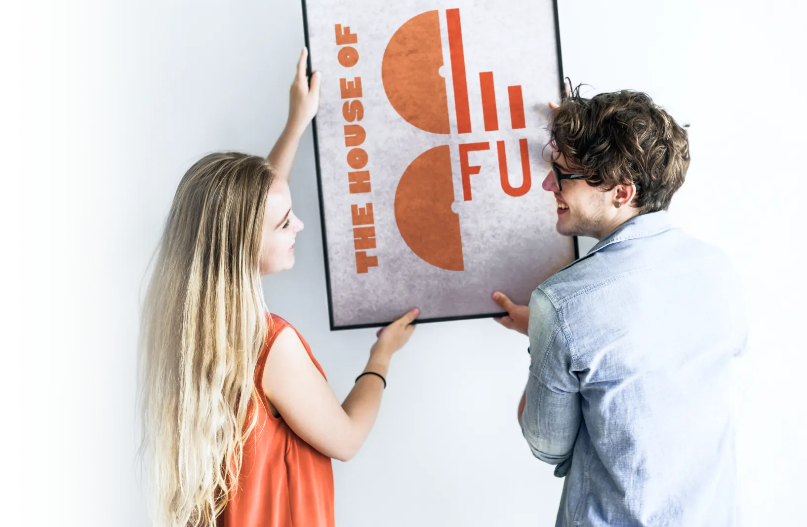 Ein lächelnder junger Mann und eine junge Frau hängen gemeinsam ein gerahmtes Poster mit der Aufschrift „THE HOUSE OF CO / FU“ in kräftigem Orange auf. Die helle, minimalistische Umgebung wirkt freundlich – eine Szene voller Teamgeist und Spaß beim Einrichten.