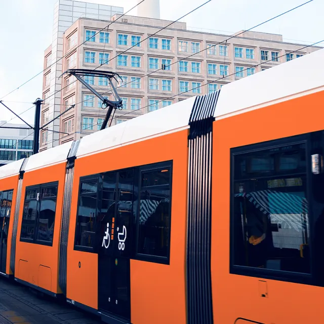 Eine leuchtend orange Straßenbahn fährt durch eine moderne Stadtstation. Im Hintergrund ist ein Gebäude mit vielen Fenstern, wahrscheinlich ein Büro- oder Verwaltungsbau. Auf der Tram sind Piktogramme für Sitzplätze für Senioren und Kinderwagen. Oberleitungen sind zu sehen, das Wetter ist klar.