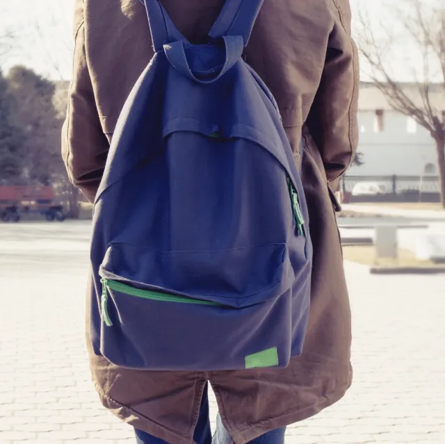 Das Bild zeigt eine Person von hinten mit braunem Mantel und großem dunkelblauen Rucksack mit grünen Reißverschlüssen. Die Person steht im Freien auf einem gepflasterten Platz, möglicherweise ein Parkweg. Kühle Witterung, kahle Bäume deuten auf Herbst oder frühen Frühling hin.