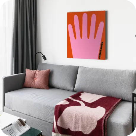 Eine stilvolle, gemütliche Ecke mit hellgrauem Sofa, weicher Decke und korallenfarbenem Kissen. Die Wandkunst – eine pinke Hand auf orange-rotem Hintergrund – setzt einen spielerischen Kontrast zur ruhigen, modernen Einrichtung.