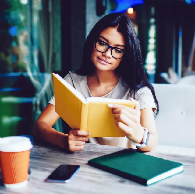 Eine junge Frau mit langen dunklen Haaren und Brille sitzt an einem Tisch und liest konzentriert ein gelbes Buch. Vor ihr stehen ein Kaffeebecher, ein Smartphone und ein grünes Notizbuch. Die Umgebung wirkt wie ein gemütliches Café oder Lernraum mit natürlichem Licht.