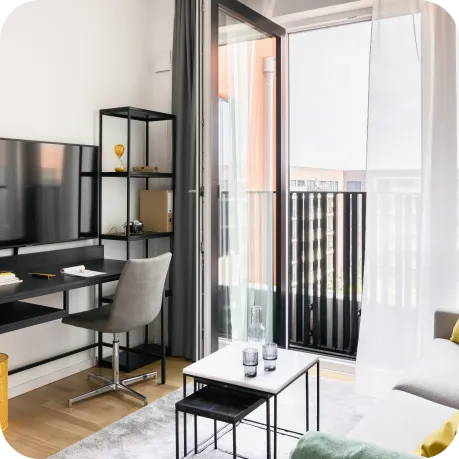 Ein heller, kompakter Wohnraum mit modernem, klarem Design. Ein Schreibtisch mit Stuhl steht gegenüber einem Wand-TV, daneben eine offene Balkontür. Sanfte Farben und Tageslicht sorgen für eine gemütliche Atmosphäre.