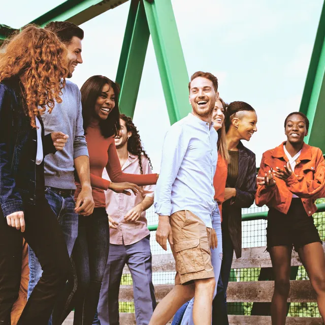 Eine vielfältige Gruppe junger Erwachsener läuft lachend über eine grüne Metallbrücke. Die Stimmung ist fröhlich und lebendig, alle unterhalten sich gut gelaunt miteinander.