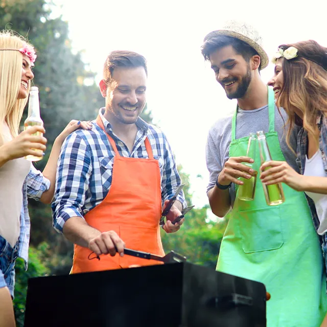 Group of four young adults enjoying a barbecue outdoors. Two men in colorful aprons are grilling while smiling and holding drinks, accompanied by two women also laughing with bottles in hand. Sunlight and trees in the background suggest a relaxed summer setting.