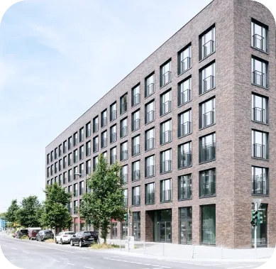 Modernes Wohngebäude mit dunkler Klinkerfassade und großen, regelmäßig angeordneten Fenstern. Der fünfstöckige Bau steht an einer ruhigen Stadtstraße mit parkenden Autos und jungen Bäumen.