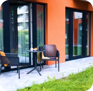 Kleine private Terrasse vor einer Wohnung mit kräftig orangefarbener Fassade. Zwei schwarze Stühle und ein runder Tisch stehen auf einer gepflasterten Fläche neben einer Rasenfläche – ein gemütlicher Außenbereich.