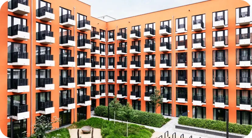 Modernes Wohngebäude mit auffälliger orangefarbener Fassade, zahlreichen identischen Balkonen und großen Fenstern. Im Innenhof befinden sich begrünte Flächen mit Bäumen, Sträuchern und einer runden Sitzecke.