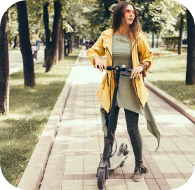 Eine junge Frau in grünem Outfit und gelber Jacke steht auf einem gepflasterten Weg im Park und hält einen E-Scooter. Umgeben von Bäumen wirkt sie selbstbewusst – ein Symbol für nachhaltige, urbane Mobilität.