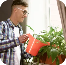 Ein junger Mann mit Brille und kariertem Hemd gießt Zimmerpflanzen auf der Fensterbank mit einer roten Gießkanne. Die Szene wirkt ruhig und wohnlich – ein Moment der Achtsamkeit oder des Urban Gardenings.