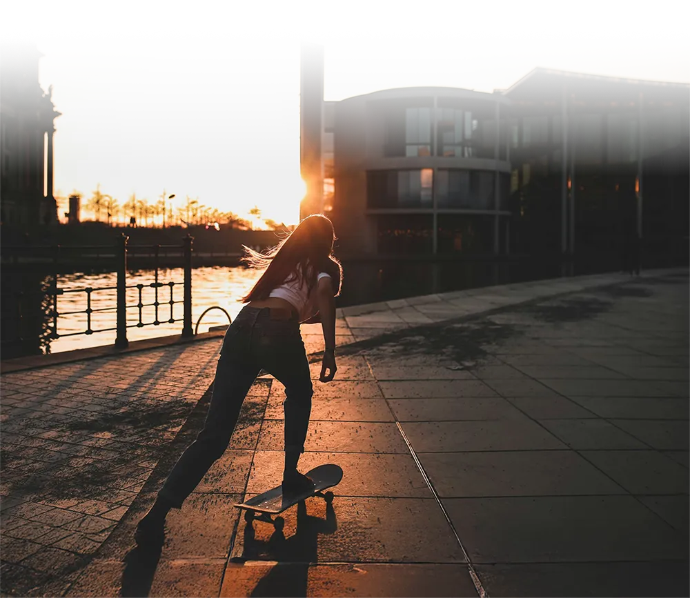 Schattenriss einer Person, die bei Sonnenuntergang mit einem Skateboard an der Uferpromenade entlangfährt. Im Hintergrund moderne Gebäude und warmes Sonnenlicht, das eine lebendige, urbane Stimmung erzeugt.