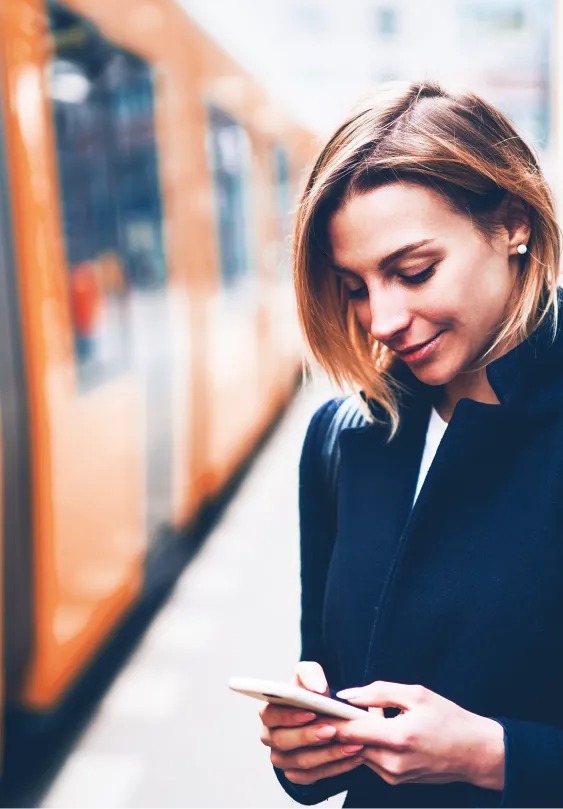 Lächelnde Frau im dunklen Mantel steht an einem Bahnsteig und blickt auf ihr Smartphone. Im Hintergrund steht eine orangefarbene Stadtbahn, leicht unscharf. Sie trägt einen schwarzen Leder-Rucksack – die städtische Umgebung lässt auf eine Pendlerfahrt oder einen Termin schließen.
