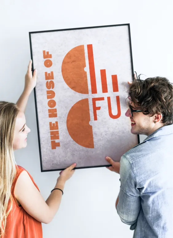 Ein lächelnder junger Mann und eine junge Frau hängen gemeinsam ein gerahmtes Poster mit der Aufschrift „THE HOUSE OF CO / FU“ in kräftigem Orange auf. Die helle, minimalistische Umgebung wirkt freundlich – eine Szene voller Teamgeist und Spaß beim Einrichten.