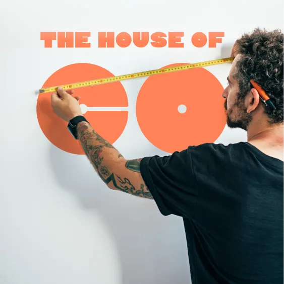 Ein Mann mit Tattoos am Arm und einem Bleistift hinter dem Ohr misst orange geometrische Formen an einer weißen Wand mit einem gelben Maßband. Über den Formen steht in fetter roter Schrift „THE HOUSE OF CO“. Das Bild vermittelt Kreativität und Präzision im Co-Living- oder Designkontext.