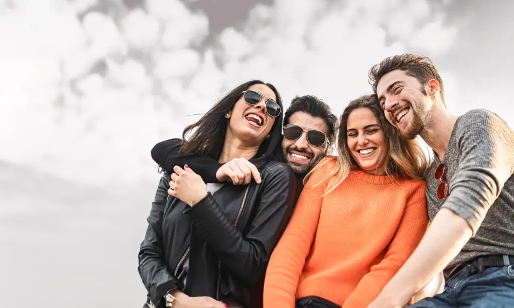 Four young friends laugh together outdoors under a cloudy sky, dressed casually with sunglasses and bright expressions. Their close, joyful pose radiates connection, fun, and carefree friendship vibes.