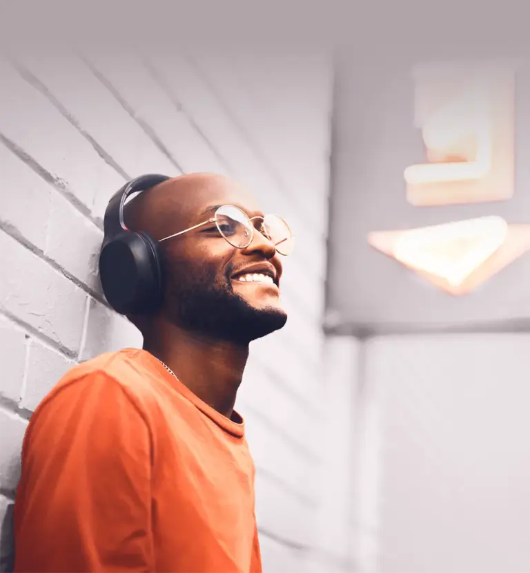 Ein lächelnder Mann lehnt an einer hellen Ziegelwand und hört Musik mit kabellosen Kopfhörern. Mit Brille und orangem Shirt strahlt er Entspannung und Zufriedenheit unter einem leuchtenden Neonschild aus.