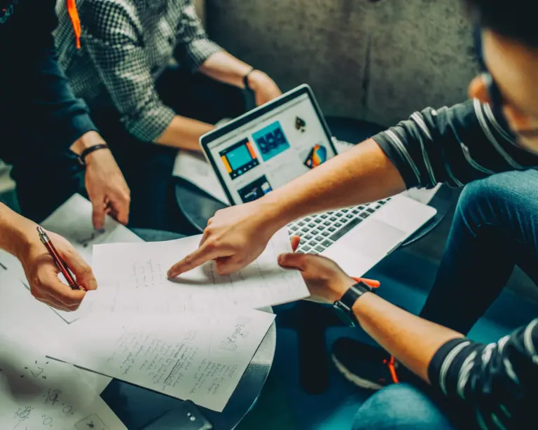 Drei Personen arbeiten intensiv an einem kreativen oder planerischen Projekt. Umgeben von handschriftlichen Notizen und einem Laptop mit Designvorschlägen, wird konzentriert diskutiert und gezeigt – eine Szene voller Teamarbeit und Ideenfindung.