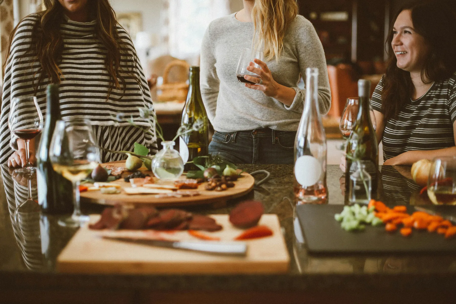 Eine Gruppe Frauen steht entspannt in einer gemütlichen Küche, trinkt Wein und unterhält sich. Auf Schneidebrettern und der Arbeitsfläche liegen Wurst, Feigen und Gemüse – es wirkt wie ein lockerer Abend unter Freunden oder gemeinsames Kochen.