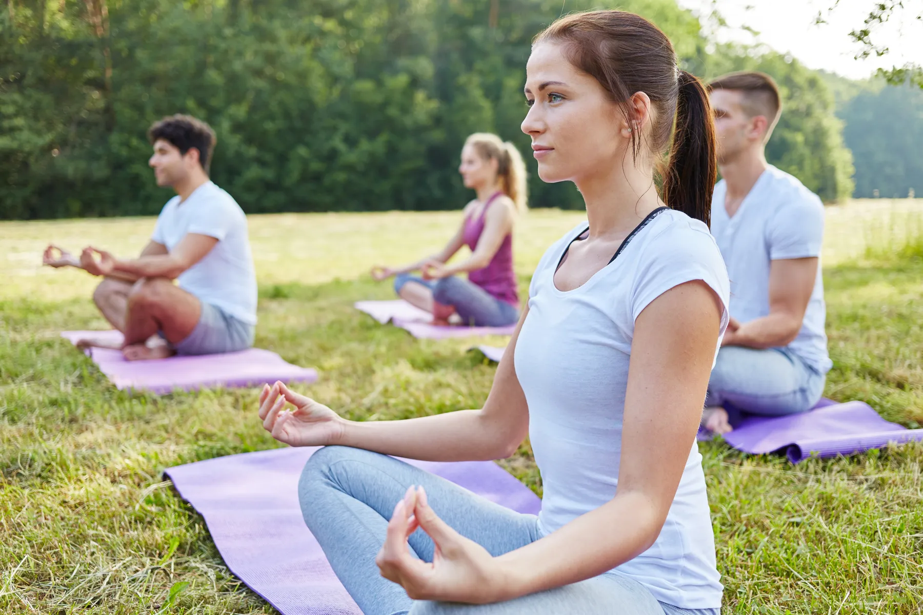 Vier Personen praktizieren entspannt Yoga und Meditation in einer ruhigen, natürlichen Umgebung im Freien. Sie sitzen im Schneidersitz auf violetten Matten, die auf einer Grasfläche ausgebreitet sind, und nehmen eine entspannte Haltung mit geschlossenen Augen oder einem sanften Blick ein, während ihre Hände ruhig auf den Knien in Meditationspose ruhen. Sanftes Sonnenlicht beleuchtet die friedliche Szene und verstärkt die Atmosphäre der Achtsamkeit, Gelassenheit und inneren Balance.