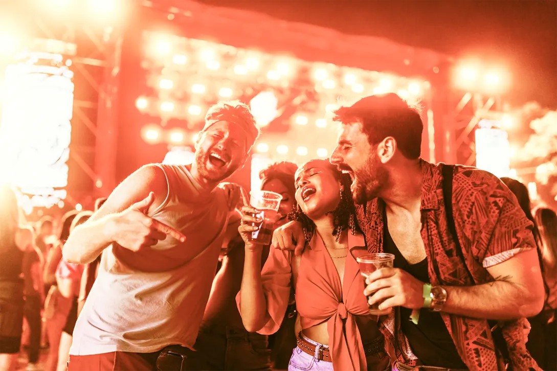 Drei junge Erwachsene feiern begeistert und singen ausgelassen bei einem lebhaften Konzert oder Festival. Sie lächeln, halten Getränke in den Händen und genießen die energiegeladene Atmosphäre, die durch die leuchtenden, bunten Bühnenlichter hervorgehoben wird. Ihre Mimik und Körpersprache vermitteln Begeisterung, Freude und unbeschwerten Spaß.
