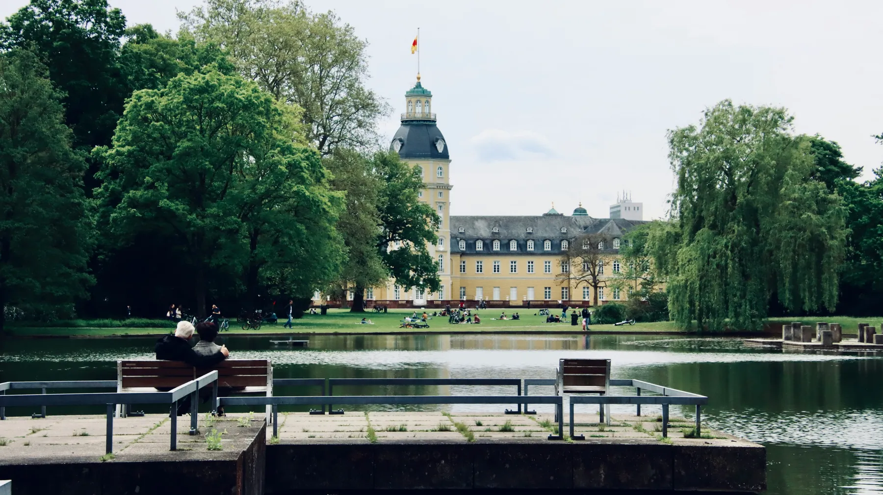 Eine friedliche Szene zweier Personen, die auf einer Bank am Teich sitzen, umgeben von üppig grünen Bäumen und einem eleganten historischen Gebäude im Hintergrund.
