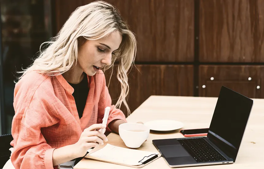 Eine konzentrierte junge Frau sitzt an einem Tisch, arbeitet am Laptop und macht Notizen auf einem Klemmbrett.