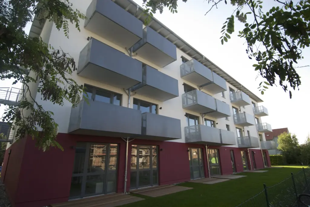 Modernes Wohngebäude mit rot-weißer Fassade, großen Fenstern und mehreren grauen Balkonen in Reihen. Der Blick erfolgt von der Gartenseite mit grünem Rasen und belaubten Ästen im Vordergrund.