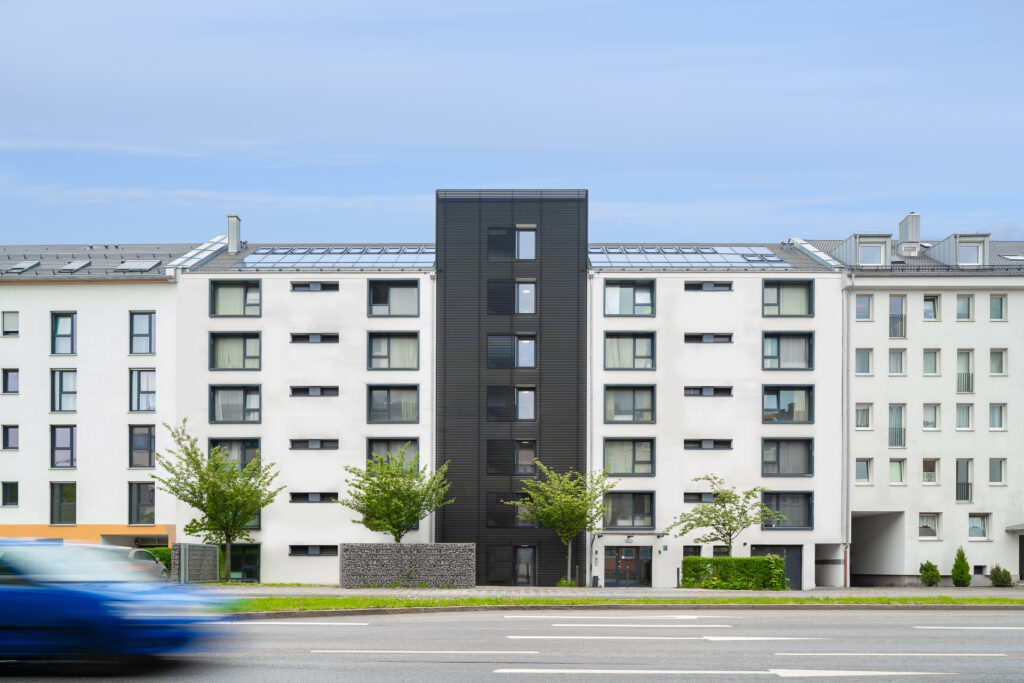 Modernes Wohngebäude mit weißer Fassade und dunklem mittleren Treppenhausbereich, an einer stark befahrenen Straße gelegen. Im Vordergrund fährt ein verschwommenes blaues Auto vorbei. Das Gebäude hat große Fenster und kleine Bäume davor.
