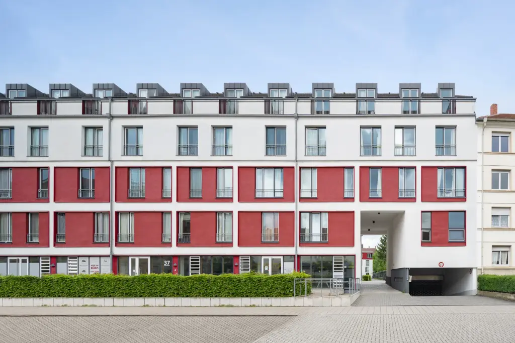 Ein modernes Wohngebäude mit einer weißen und roten Fassade, mehreren Fensterreihen und kleinen Balkonen. Der Eingang führt durch einen Torbogen in der Mitte des Gebäudes, davor befindet sich ein gepflasterter Bereich mit grünen Hecken.