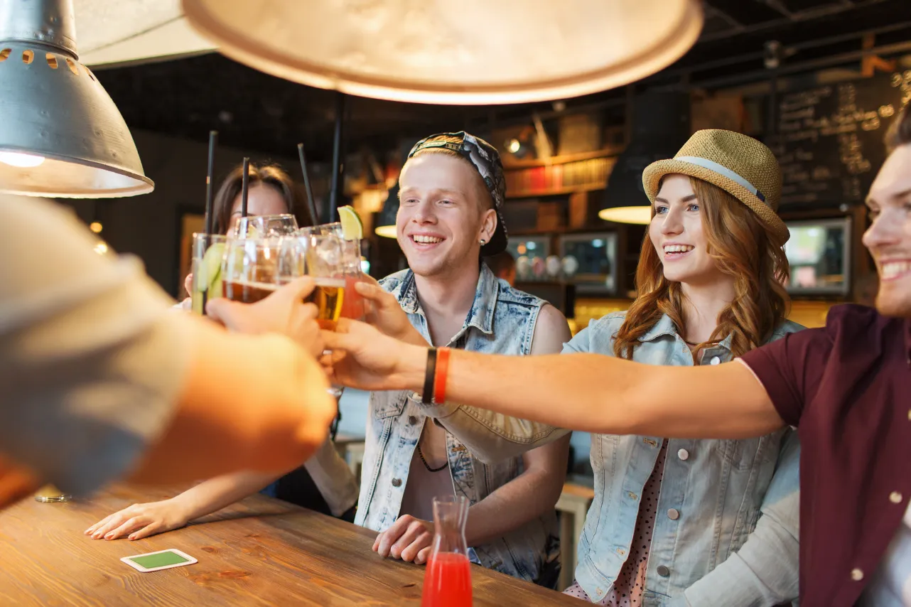 A group of cheerful young friends clinking glasses in celebration at a cozy, warmly-lit bar.