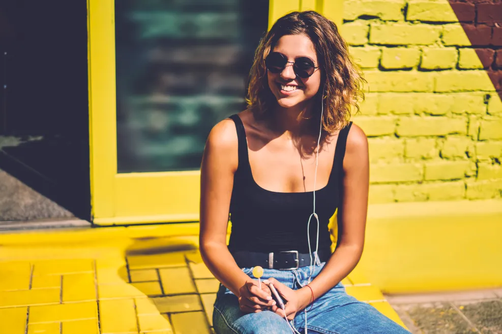 Young smiling woman wearing sunglasses and earphones, sitting casually in front of a brightly colored yellow and green wall.