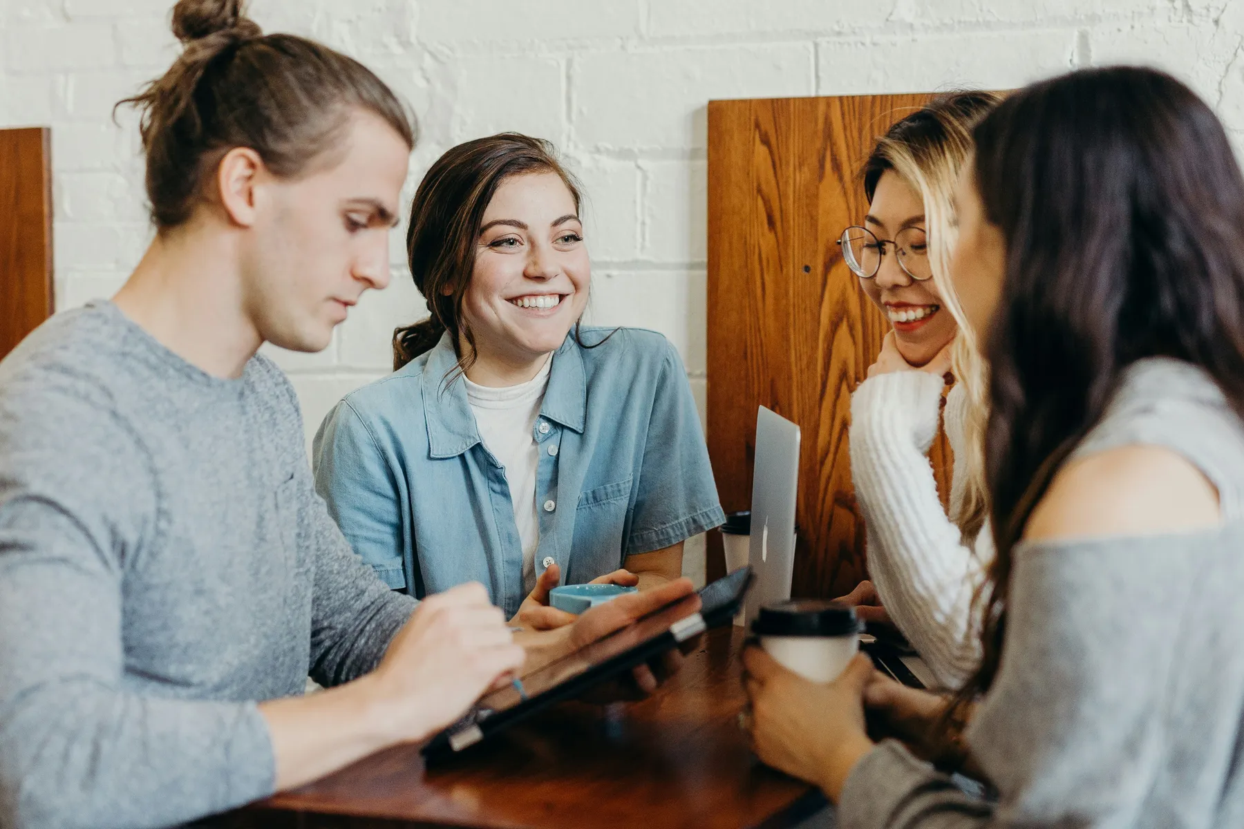 A group of four young adults smiling, talking, and working together at a wooden table with laptops and coffee cups.