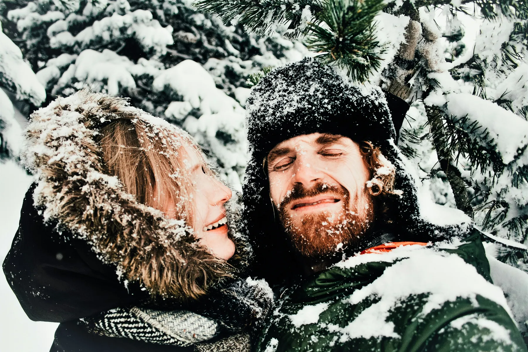 Glückliches Paar macht ein Selfie im verschneiten Wald, ihre Gesichter sind mit Schnee bedeckt, warm eingepackt in Winterkleidung.