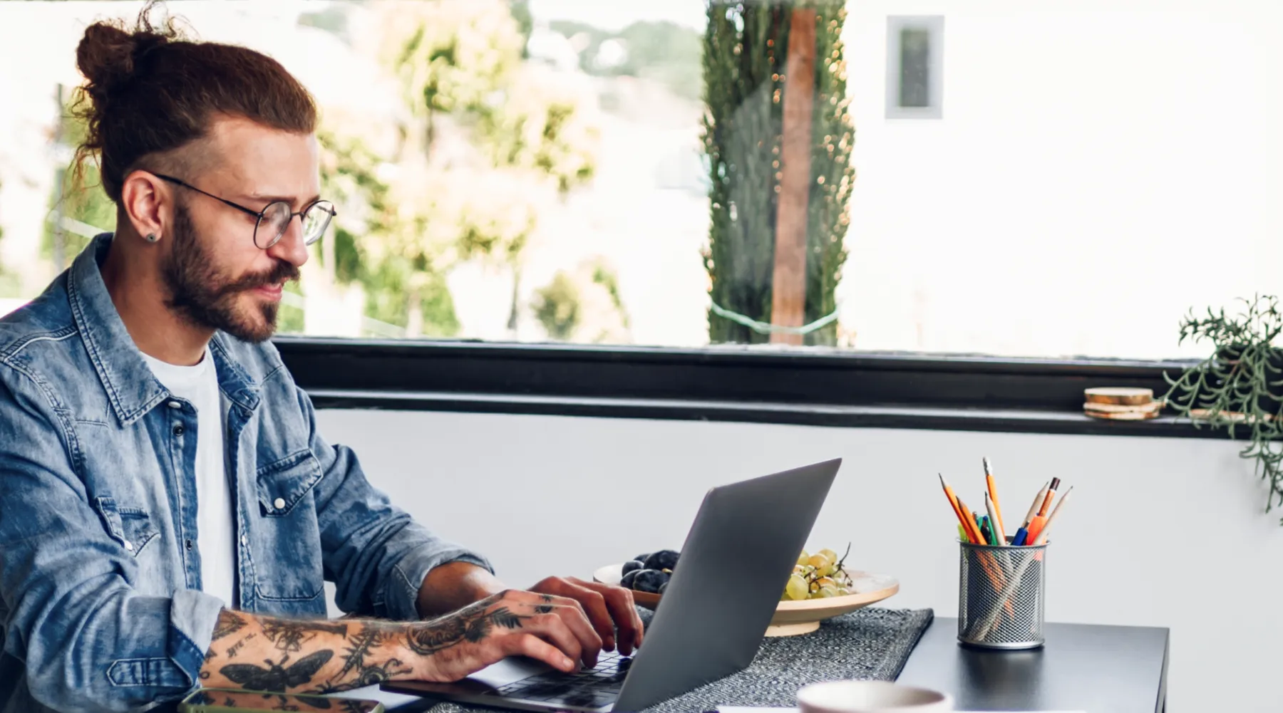 Ein junger Mann mit Tätowierungen und Brille arbeitet an einem Laptop in einem hellen, modernen Arbeitsbereich.
