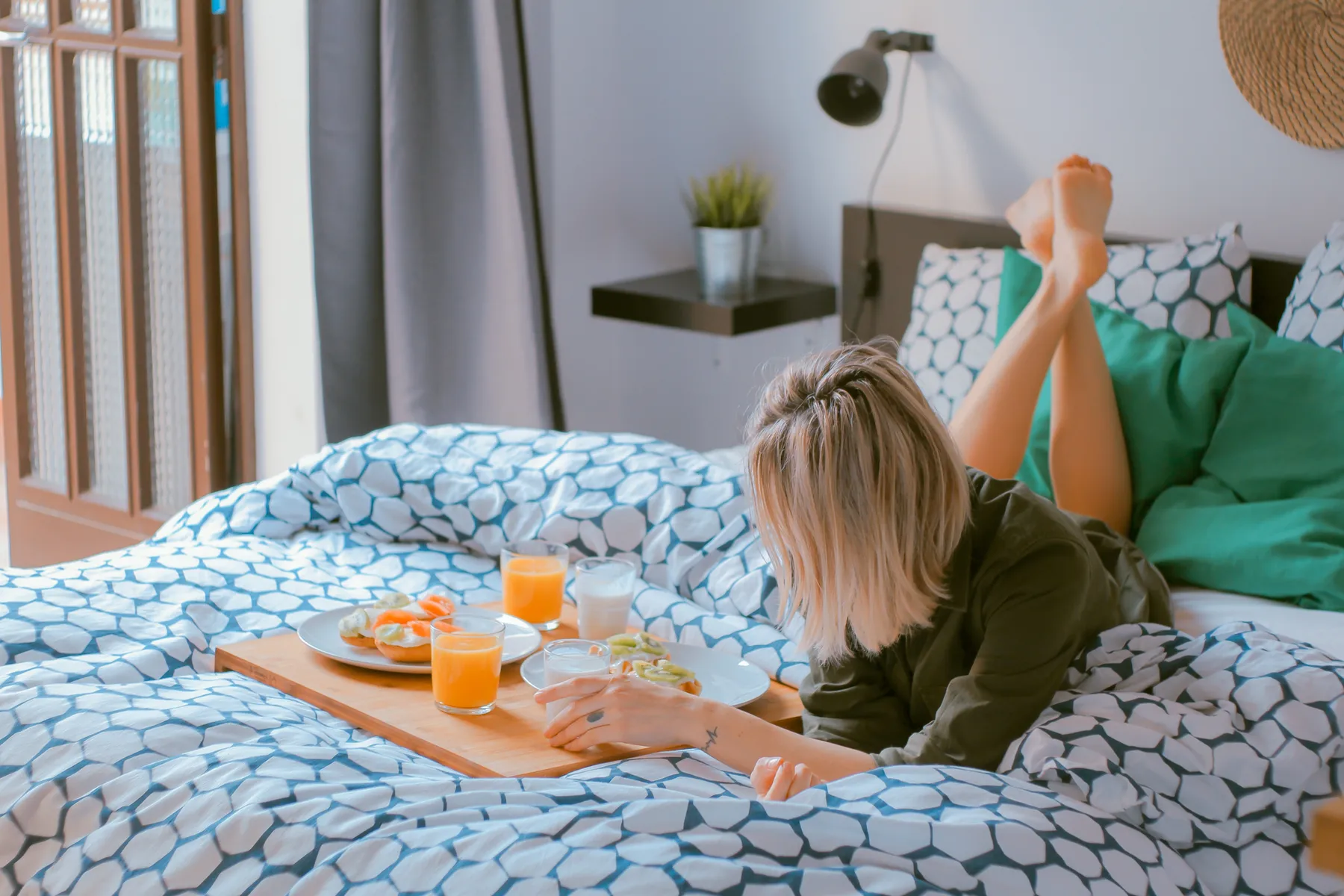 A young woman lounges comfortably on a cozy, patterned bed with her legs up, enjoying a breakfast tray with juice, milk, fruit, and toast. Soft light and fresh tones evoke a peaceful, slow morning at home.