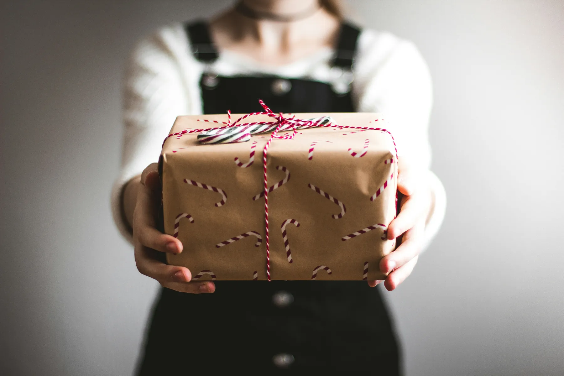 Eine Person in schwarzem Latzrock hält ein liebevoll verpacktes Geschenk mit Zuckerstangenmotiv und rotem Band entgegen. Der neutrale Hintergrund lenkt den Blick ganz auf die herzliche Geste des Schenkens.