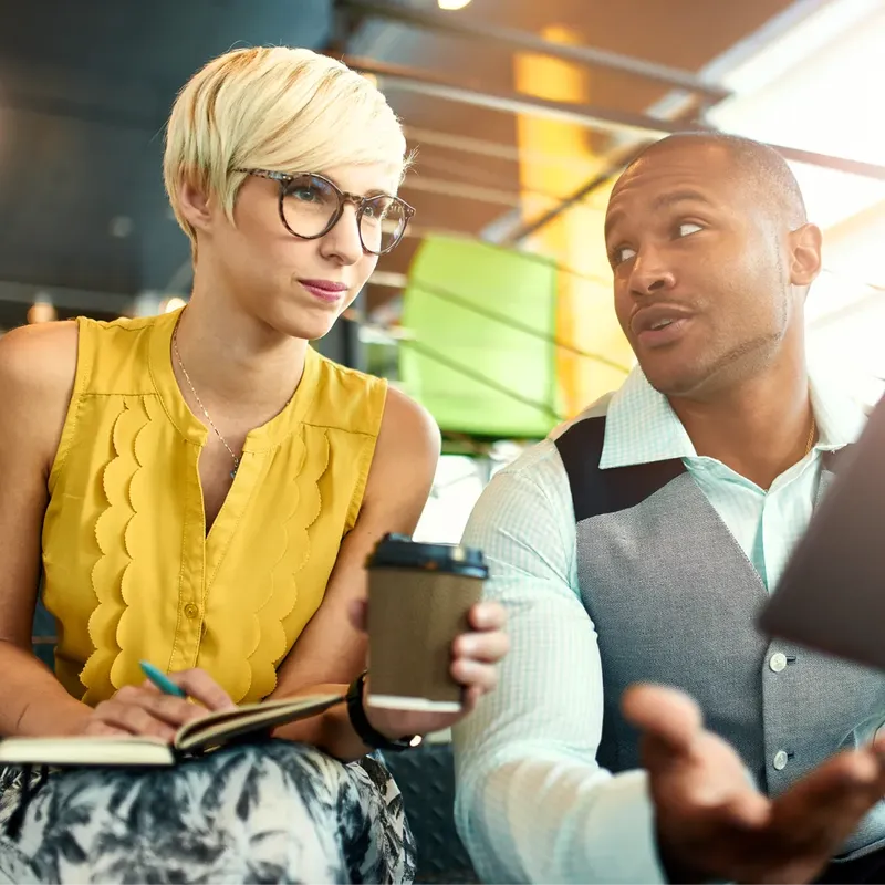 Ein Mann und eine Frau unterhalten sich in einem modernen Büro oder Co-Working-Space. Die Frau mit Brille und gelber Bluse macht Notizen in ein Notizbuch und hält einen Kaffeebecher. Der Mann trägt ein Hemd mit Weste, gestikuliert mit der Hand und hält ein Tablet.