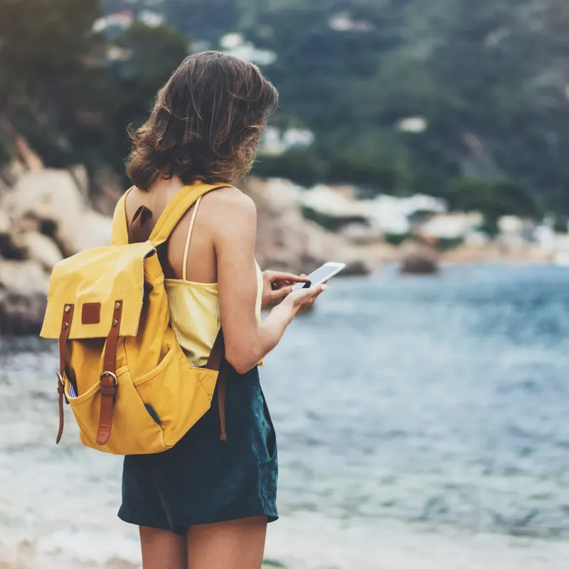 Eine junge Frau mit einem gelben Rucksack steht an einer felsigen Küste und schaut auf ihr Smartphone. Sie trägt ein leichtes Oberteil und Shorts. Im Hintergrund sind verschwommene Berge und Küstengebäude zu sehen.