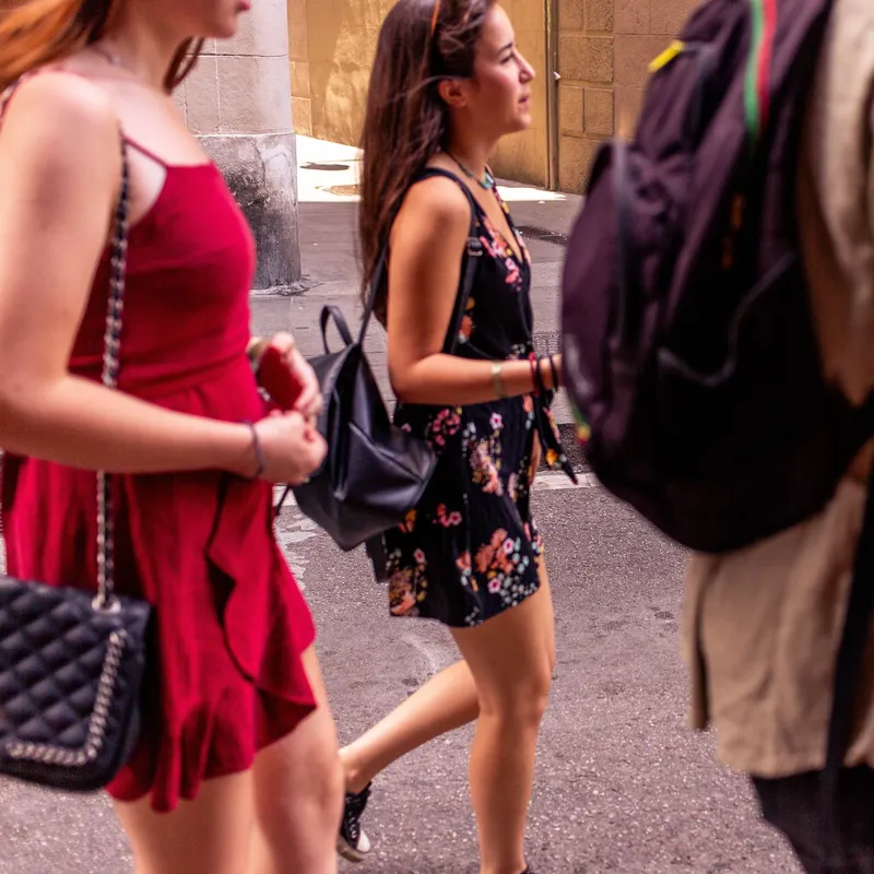 Eine Gruppe junger Menschen geht bei warmem Wetter eine Stadtstraße entlang. Zwei Frauen in Sommerkleidern und ein Mann mit Rucksack sind im Vordergrund zu sehen, während eine weitere Person in dunkler Kleidung mit einer Kamera auf der gegenüberliegenden Straßenseite läuft. Die Umgebung wirkt urban mit Steinbauten und Pflastersteinen.