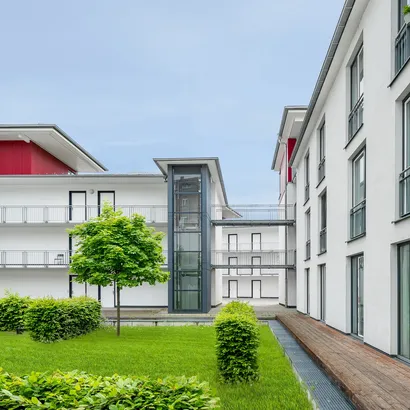 Ein Innenhof eines modernen Wohnkomplexes mit gepflegtem grünen Rasen, Büschen und einem kleinen Baum. Die Gebäude sind weiß mit roten Akzenten und großen Fenstern. In der Mitte befindet sich ein gläserner Aufzugsschacht.