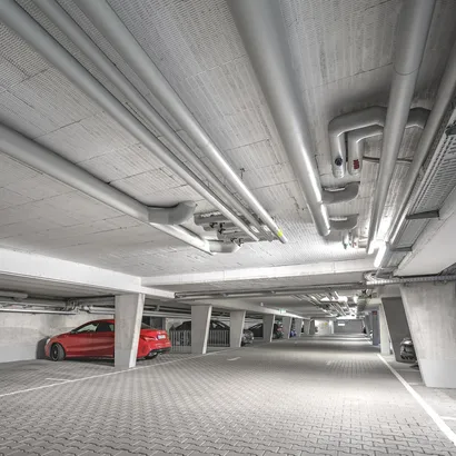 Ein Tiefgaragenparkplatz mit Betonboden und -decke. Große Industrie-Rohre verlaufen über die Decke. Auf der linken Seite ist ein rotes Auto geparkt, weitere Fahrzeuge sind in der Tiefe zu sehen. Die Beleuchtung ist hell und gleichmäßig verteilt.