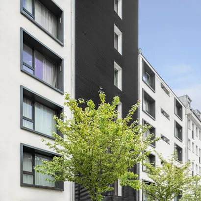 Modernes Wohngebäude mit einer Fassade aus weißen und dunkelgrauen Paneelen. Das Gebäude verfügt über große Fenster und ist von grünen Bäumen gesäumt, unter einem klaren blauen Himmel.