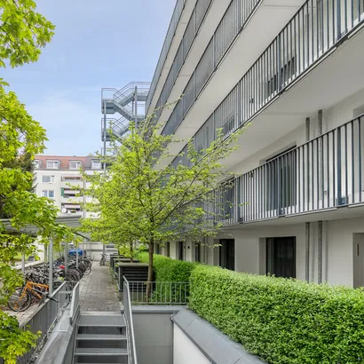 Modernes Wohngebäude mit Balkonen und einem angelegten Innenhof. Der Innenhof ist mit grünen Hecken, jungen Bäumen und einer Reihe geparkter Fahrräder entlang eines Gehwegs ausgestattet