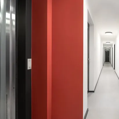 Moderner Innenflur eines Wohngebäudes mit weißen Wänden, schwarzen Leisten und einer auffälligen roten Akzentwand in der Nähe eines Aufzugs.