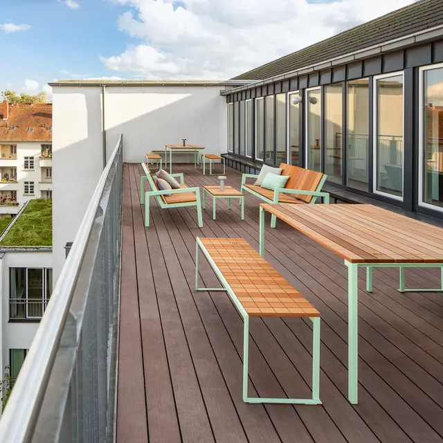 Eine Dachterrasse mit Holzdeck und modernen Gartenmöbeln. Die Möbel haben Holzflächen mit mintgrünen Metallrahmen, darunter Bänke, Tische und Loungesessel mit pastellfarbenen Kissen. Die Terrasse ist von Glasgeländern umgeben und grenzt an ein Gebäude mit großen Fenstern. Der Himmel ist teils bewölkt und sorgt für eine helle, angenehme Atmosphäre.