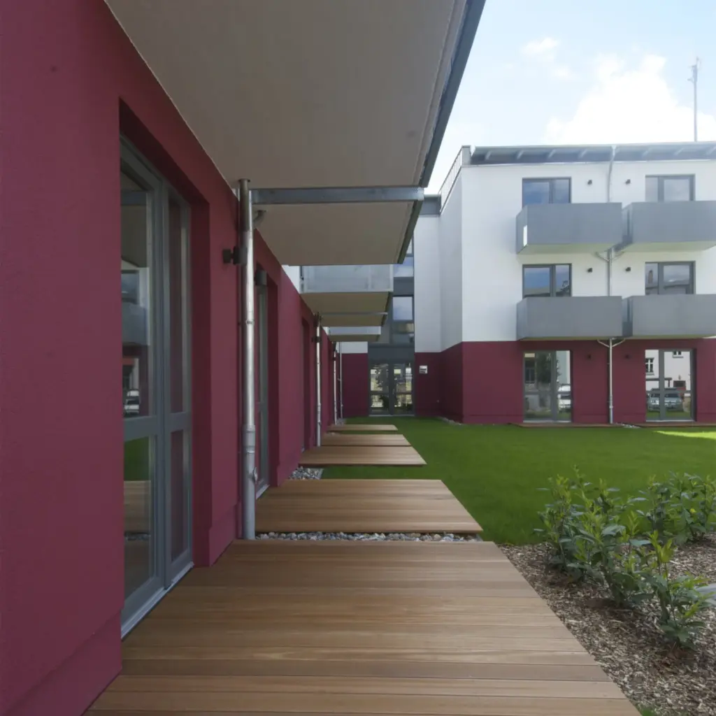 Blick auf ein modernes Apartmenthaus mit weinroter und weißer Fassade auf Erdgeschosshöhe. Holzterrassen vor jeder Einheit, daneben ein gepflegter Rasen und junge Pflanzen.