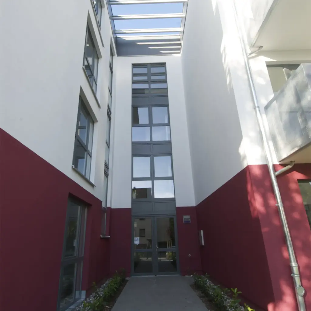 Blick in den Innenhof eines modernen Wohngebäudes mit klaren architektonischen Linien. Die Fassade ist in Weiß und Dunkelrot gehalten, mit großen Fenstern und einem Glasdach darüber, das Tageslicht in den Eingangsbereich lässt.