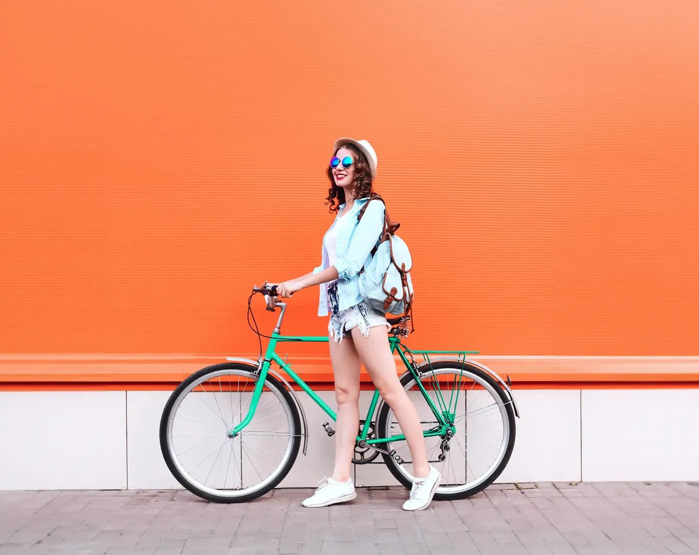 Junge Frau mit Sonnenbrille, Hut und Rucksack steht mit einem grünen Fahrrad vor einer leuchtend orangefarbenen Wand.